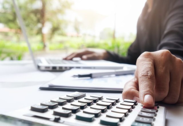 Main utilisant une calculatrice devant un ordinateur portable et des documents comptables