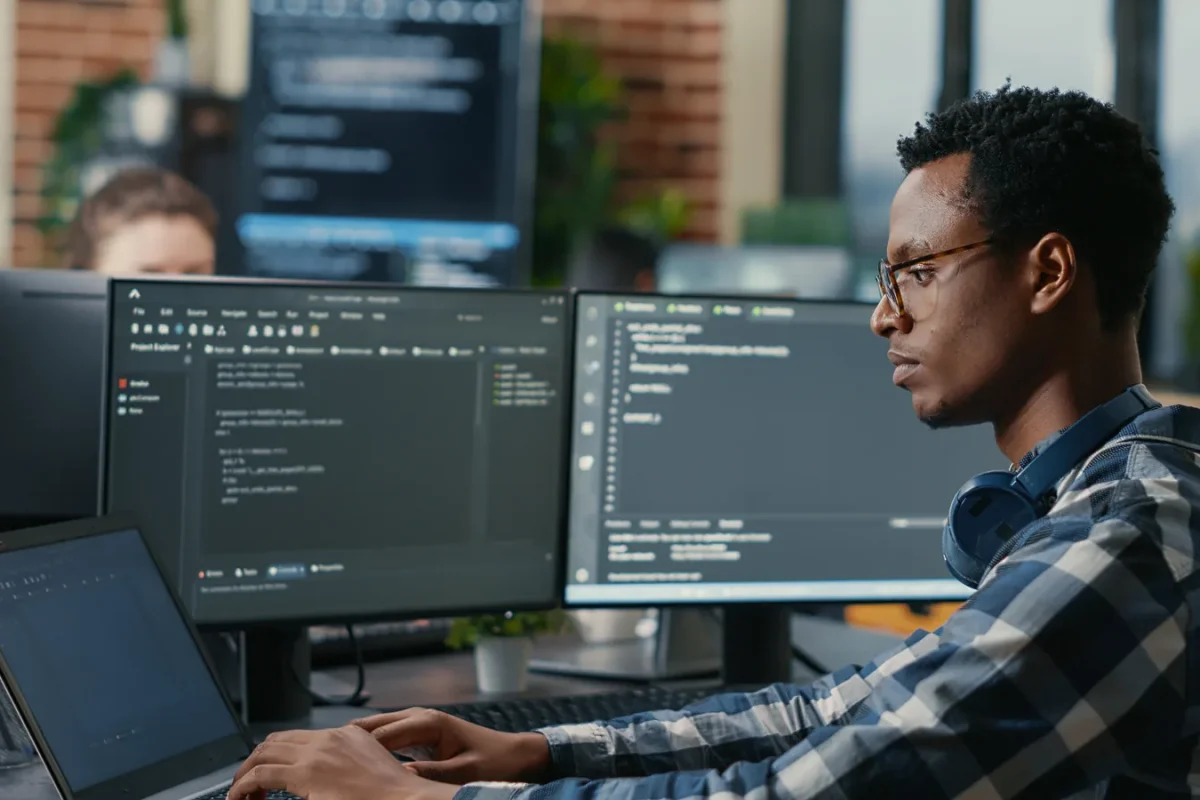 Développeur à lunettes travaillant sur un ordinateur portable devant deux écrans affichant du code