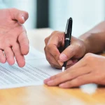 Une main désigne un paragraphe d’un contrat pendant qu’une autre personne le signe au stylo