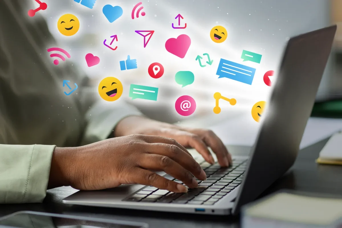 Mains tapant sur le clavier d’un ordinateur portable d’où s’échappent des icônes de réseaux sociaux