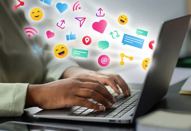 Mains tapant sur le clavier d’un ordinateur portable d’où s’échappent des icônes de réseaux sociaux