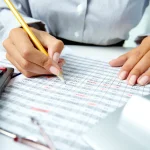Personne vérifiant au crayon un tableau de chiffres imprimé, à côté de calculatrices