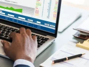 Mains sur le clavier d’un ordinateur portable affichant des tableaux de chiffres, à côté d’une facture posée sur un porte-bloc