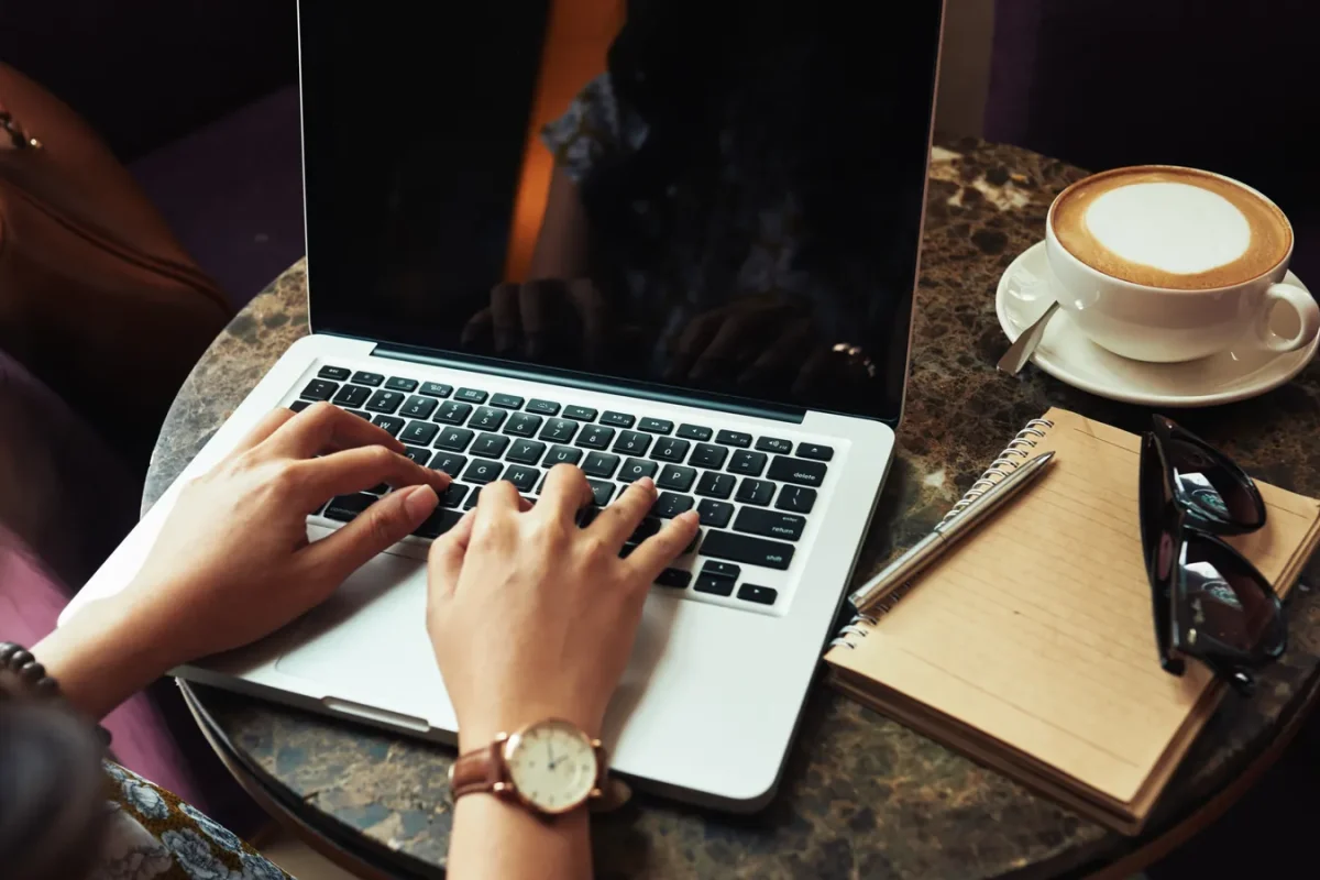 Personne rédigeant un texte sur un ordinateur portable, carnet et café posés sur la table