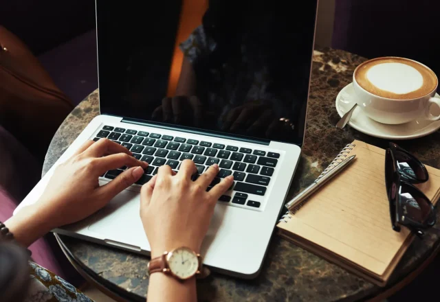 Personne rédigeant un texte sur un ordinateur portable, carnet et café posés sur la table