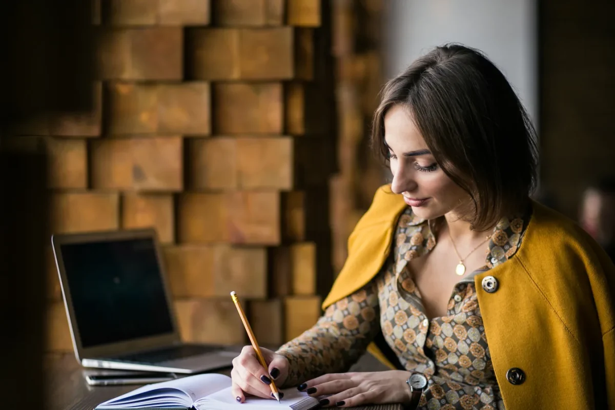Femme assise à une table écrivant au crayon dans un carnet, un ordinateur portable ouvert à côté d’elle