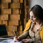 Femme assise à une table écrivant au crayon dans un carnet, un ordinateur portable ouvert à côté d’elle
