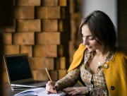 Femme assise à une table écrivant au crayon dans un carnet, un ordinateur portable ouvert à côté d’elle