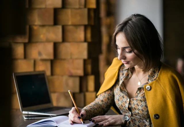 Femme assise à une table écrivant au crayon dans un carnet, un ordinateur portable ouvert à côté d’elle