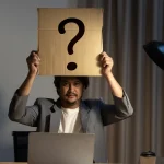 Homme assis devant son ordinateur portable, tenant au-dessus de sa tête une pancarte en carton marquée d’un point d’interrogation