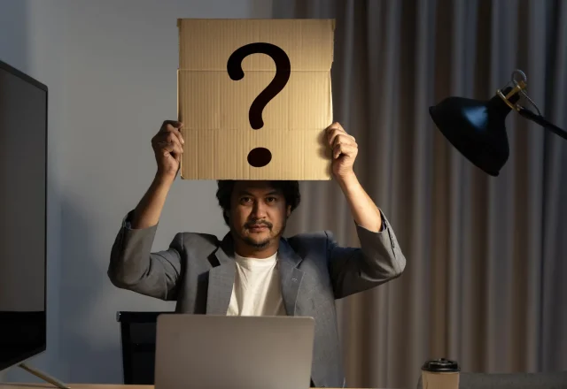 Homme assis devant son ordinateur portable, tenant au-dessus de sa tête une pancarte en carton marquée d’un point d’interrogation