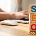 Cubes en bois formant le mot SEO avec l'année 2027, devant une personne travaillant sur un ordinateur portable