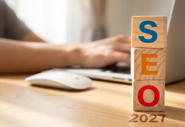 Cubes en bois formant le mot SEO avec l'année 2027, devant une personne travaillant sur un ordinateur portable