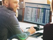 Homme barbu échangeant avec une collègue devant un écran affichant un tableur de données comptables