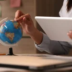 Mains d’une personne faisant tourner un petit globe terrestre tout en consultant une tablette à son bureau