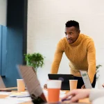 Homme debout penché sur une table de réunion face à des collègues équipés d'ordinateurs portables