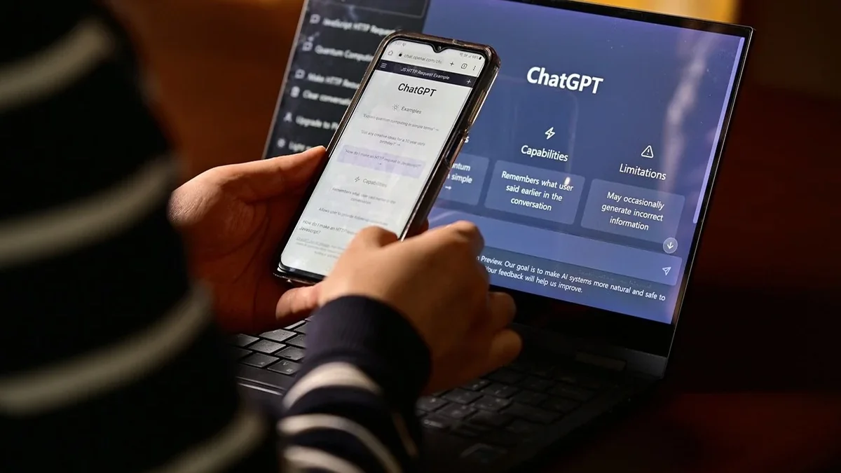 Personne consultant ChatGPT sur un smartphone devant un ordinateur portable